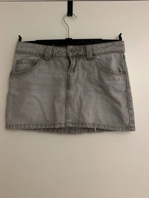 Grå jeanskjol med fickor - Säljer en grå jeanskjol i kort modell med klassiska fem fickor, bälteshällor och knappstängning fram. Kjolen har en rak passform och är perfekt för dig som gillar en enkel och stilren look.
