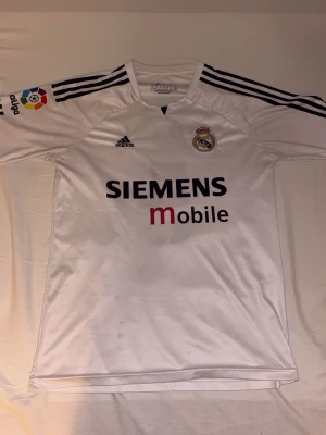 Vit Real Madrid matchtröja Adidas S - Klassisk vit Real Madrid matchtröja från Adidas med svarta detaljer och klubbmärke på bröstet. Siemens Mobile-logga på framsidan och La Liga-märke på ärmen. Tillverkad i lätt och ventilerande material, perfekt för fotboll eller träning.