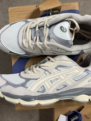 Asics vita och grå sneakers - Snygga sneakers från Asics i vitt och ljusgrått med mesh- och mockadetaljer. Skorna har en sportig design med tydliga Asics-loggor på sidan och tungan. Sulan är tjock och ger bra stöd, perfekt för en trendig streetwear-look.