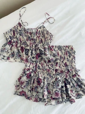 Blommigt tvådelat set med volanger - Säljer ett superfint tvådelat set med blommönster i vitt, lila och svart. Setet består av ett linne med tunna axelband som knyts och volang samt en matchande kjol med smock i midjan och dubbla volanglager. Materialet är tunt och luftigt💜💜