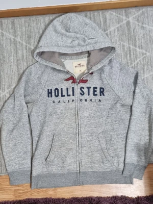 Grå zip hoodie från Hollister - Snygg gråmelerad hoodie från Hollister med dragkedja, huva och fickor framtill. Stor Hollister-logga och text i mörkblått samt röd fågel på bröstet. Mjuk bomullsblandning och ribbade muddar vid ärmar och nederkant. Perfekt för chill dagar.