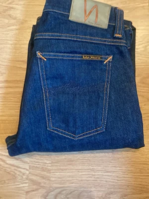 Mörkblå slim fit jeans från Nudie Jeans - Säljer ett par mörkblå jeans från Nudie Jeans w28 med klassisk femficksmodell och kontrastsömmar. Jeansen har smal passform och är tillverkade i slitstark denim. Perfekta för dig som gillar en clean och tidlös look.