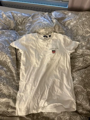 Vit t-shirt från Gant med logga - Säljer en klassisk vit t-shirt från Gant med rund hals och korta ärmar. På bröstet finns Gant-loggan och ett broderat sköldmärke i rött, vitt och blått. Tillverkad i mjuk bomull som är skön att bära. Perfekt till jeans eller shorts.