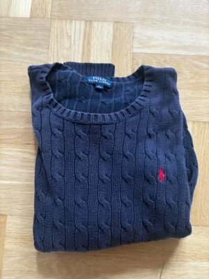 Mörkblå stickad tröja Polo Ralph Lauren - Mörkblå kabelstickad tröja från Polo Ralph Lauren med rund halsringning och röd broderad logga på bröstet. Tröjan har långa ärmar och ribbade muddar vid ärmslut och nederkant. Perfekt för en stilren och klassisk look.