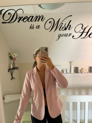 Ljusrosa kavaj med klassisk passform - Snygg ljusrosa kavaj med klassisk krage och två fickor framtill. Figurnära modell som ger en stilren look. Perfekt att matcha med både jeans och kostymbyxor. Materialet ser ut att vara mjukt och bekvämt, troligen bomulls- eller polyestermix.