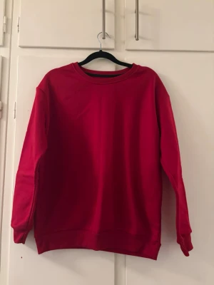 Röd basic sweatshirt - En enkel röd sweatshirt med rund halsringning och långa ärmar. Tröjan har ribbade muddar vid ärmslut och nederkant. Perfekt för en avslappnad och stilren look. Materialet ser ut att vara mjukt och bekvämt, troligen bomull eller bomullsblandning.
