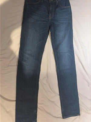 Blå raka jeans med klassisk passform - Säljer ett par blå jeans med rak passform och fem fickor. Jeansen har normal midja, klassiska sömmar och en läderpatch bak i linningen. Materialet är denim i bomull och färgen är en snygg mellanblå tvätt. Perfekta till en avslappnad stil.