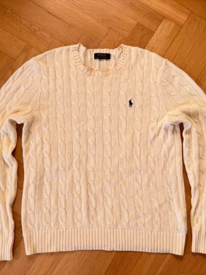 Polo Ralph Lauren kabelstickad Tröja Cream M - Kabelstickad tröja i krämvitt från Polo Ralph Lauren med rund halsringning och broderad marinblå logga på bröstet. Tröjan har ribbade muddar vid ärmslut och nederkant. Perfekt för en klassisk och stilren look.