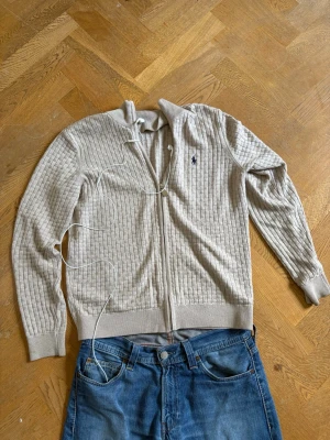 Ljusgrå ribbad ziptröja Polo Ralph Lauren - Ljusgrå ribbstickad ziptröja från Polo Ralph Lauren med hög krage och dragkedja framtill. Tröjan har långa ärmar, ribbade muddar och en liten mörkblå logga broderad på bröstet. Perfekt för en avslappnad men stilren look. Skriv vid minsta fundering och priset är inte hugget i sten 