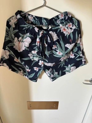 Blommiga shorts med snörning - Säljer ett par mörkblå shorts med blommigt mönster och gröna blad. Shortsen har snörning i midjan för justerbar passform och är perfekta för sommaren. Lätta och sköna att bära, passar till många olika stilar.(Badshorts)
