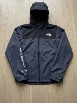 Svart vindjacka från The North Face - Snygg svart vindjacka med huva från The North Face. Jackan har dragkedja framtill, logga på bröstet och text på ena ärmen. Tillverkad i lätt och tåligt material, perfekt för blåsiga dagar. Elastiska muddar vid ärmsluten och justerbar huva.