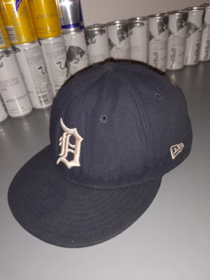 Svart New Era keps med D-logga - Snygg svart keps från New Era med en broderad vit D-logga framtill. Klassisk modell med böjd skärm och ventilerande hål upptill. Perfekt för dig som vill ha en stilren och sportig look.