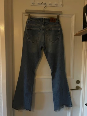 Blå bootcut jeans från Lee - Säljer ett par klassiska blå bootcut jeans från Lee med råa, fransiga benslut och femficksmodell. Jeansen har hög midja och är tillverkade i denim med en snygg tvätt. Perfekta för en avslappnad och trendig look.