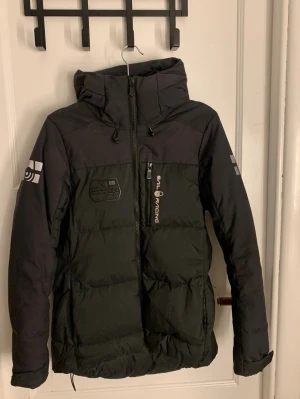 Svart dunjacka från Sail Racing - Snygg svart dunjacka från Sail Racing med huva och dragkedja framtill. Jackan har logotyp på bröstet och ärmen samt en bröstficka med dragkedja. Tillverkad i slitstarkt material och har en sportig look med detaljer i grått. Väldigt varm perfekt nu för vintern 
