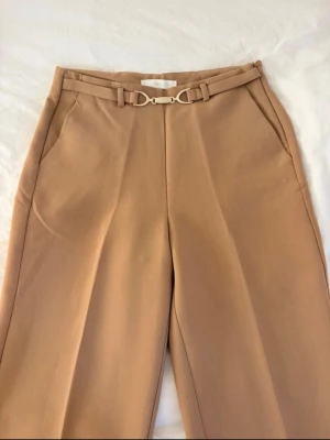 Beige kostymbyxa med bälte från Mango - Stilrena beige kostymbyxor från Mango med hög midja och raka ben. Byxorna har ett matchande tygbälte med guldfärgat spänne och diskreta sidofickor. Perfekta för en dressad look och tillverkade i ett mjukt, vävt material.