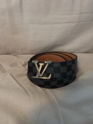 Lv bälte - Snyggt svart bälte från Louis Vuitton med klassiskt rutigt mönster och stor LV-logga i silver på spännet. Bältet har en exklusiv känsla och är tillverkat i skinn med detaljer i silverfärgad metall. Perfekt accessoar för att lyfta din outfit.