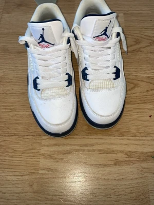 Vita och marinblå Air Jordan 4 sneakers - Snygga Air Jordan 4 sneakers i vitt läder med marinblå och grå detaljer. Skorna har meshpaneler, plastdetaljer och klassisk Jumpman-logga på plös och häl. Perfekta för dig som vill sticka ut med en ikonisk och sportig look.