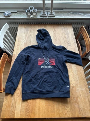 Helt Hansen hoodie med Stockholm-tryck - Säljer en mörkblå hoodie med huva och dragsko. På bröstet finns ett tryck med texten 'STOCKHOLM' och grafiskt motiv i vitt och rött. Hoodien har en stor magficka och ribbade muddar vid ärmslut och nederkant. Perfekt för dig som gillar enkel och stilren streetwear.