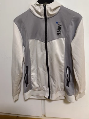 Nike hoodie i vitt och grått - Nike hoodie med dragkedja i vitt och ljusgrått. Hoodien har två svarta fickor med dragkedja framtill, svart Nike-logga på bröstet och en huva. Tillverkad i ett glansigt, mjukt material som känns sportigt och bekvämt.