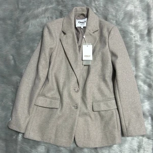 ONLY jacka onlEira-Wembley L/S Ovs Blazer - Stilren beige kavaj från ONLY med klassisk krage, två knappar framtill och två fickor med lock. Kavajen har en mjuk, slät yta och är helfodrad för extra komfort. Perfekt för en trendig och uppklädd look.