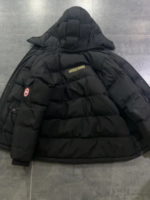 Svart dunjacka från Canada Goose - Säljer en svart dunjacka från Canada Goose med huva och klassisk patch på ärmen. Jackan har quiltad design och en markerad logotyp på ryggen. Perfekt för kalla vinterdagar och riktigt varm tack vare dunfyllningen.