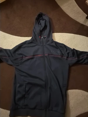 Mörkblå hoodie från Hugo Boss - Snygg mörkblå hoodie från Hugo Boss med dragkedja och huva. Diskret BOSS-logga på bröstet och en tunn röd och vit rand längs bröstet. Perfekt för en chill och stilren look. Mjukt material och två fickor framtill.