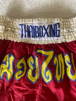 Röda thaiboxningsshorts - Säljer ett par röda thaiboxningsshorts Shortsen har gula och blå detaljer samt thailändsk text på framsidan. Perfekta för träning inom kampsport.