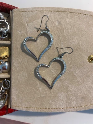 Silvriga hjärtformade örhängen - Säljer ett par stora, silvriga örhängen i form av hjärtan. Örhängena är dekorerade med gnistrande stenar längs kanten som ger en extra touch av bling. Perfekta för dig som vill sticka ut och addera lite glamour till din stil.