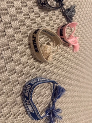 Dior J'Adior broderade armband - Fyra snygga broderade armband från Dior J'Adior-kollektionen i färgerna blå, beige, rosa och svart. Armbanden har tofsar i ändarna och texten 'J'ADIOR' broderad på bandet. Perfekt accessoar för att lyfta din outfit.