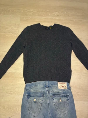 Grå kabelstickad tröja Polo Ralph Lauren - Mysig grå kabelstickad tröja från Polo Ralph Lauren med rund halsringning och diskret broderad logga på bröstet. Tröjan har långa ärmar och ribbade muddar vid ärmslut och nederkant. Perfekt för kyliga dagar och enkel att matcha.