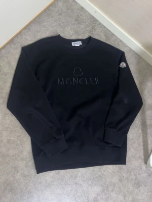 Svart sweatshirt från Moncler - Svart sweatshirt från Moncler med diskret broderad logga på bröstet och klassisk rund hals. Tröjan har ribbade muddar vid ärmslut och nederkant samt Moncler-märke på ena ärmen. Perfekt för en clean och stilren look.