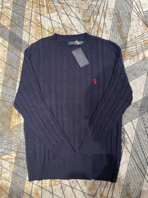 Mörkblå kabelstickad tröja Polo - Snygg mörkblå kabelstickad tröja från Polo Ralph Lauren med rund halsringning och den klassiska röda logotypen broderad på bröstet. Tröjan har långa ärmar och ribbade muddar vid ärmslut och nederkant. Perfekt för en stilren och tidlös look.