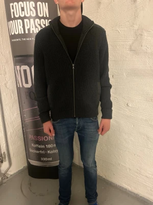 Mörkgrön stickad tröja  - Mörkgrå stickad hoodie med dragkedja framtill och ribbad struktur. Tröjan har huva, långa ärmar och muddar vid ärmslut och nederkant. Perfekt för lager på lager och en avslappnad stil.
