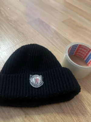 Svart ribbstickad mössa  - Size xs. Säljer en svart ribbstickad mössa med uppvikt kant och en rund patch framtill. Mössan har en klassisk, enkel design och är perfekt för att hålla värmen under kalla dagar. Materialet ser ut att vara mjukt och stretchigt, vilket gör den bekväm att bära.