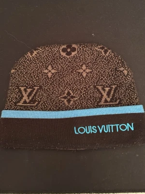 Brun mössa med blå kant - Snygg brun mössa  med klassiskt monogrammönster och ljusblå kant. Märkets logga är broderad i blått framtill. Perfekt accessoar för dig som gillar exklusiv streetstyle och vill sticka ut med en ikonisk design.