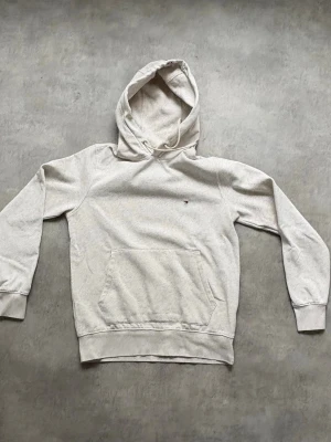 Tommy Hillfinger hoodie - Snygg beige hoodie med klassisk huva och stor magficka framtill. Liten broderad logga på bröstet och ribbade muddar vid ärmar och nederkant. Perfekt för en avslappnad och stilren look.