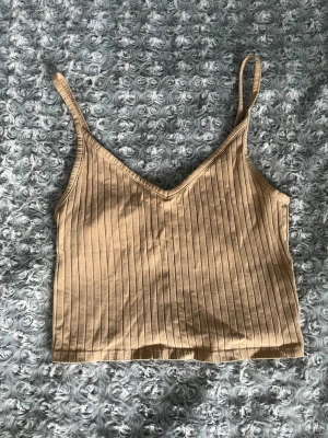 Beige ribbad linnetopp - Säljer en beige ribbad linnetopp med smala axelband och v-ringad hals. Toppen är croppad och har en enkel, stilren design som passar perfekt till sommaren. Materialet känns mjukt och bekvämt mot huden.