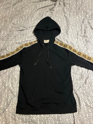 Svart hoodie med beige Gucci-detaljer - Snygg svart hoodie från Gucci med huva och dragsnören. Ärmarna har beige band med klassiska GG-loggor längs hela axeln och armen. Hoodien har känguruficka framtill och ribbade muddar. Perfekt för dig som vill ha en exklusiv och trendig look.