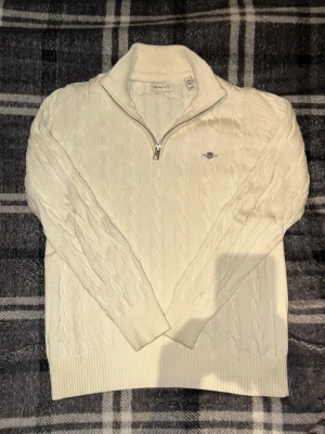 Vit stickad halv zip tröja från Gant - Vit kabelstickad tröja från Gant med halv dragkedja och ståkrage. Tröjan har ett diskret Gant-logo broderat på bröstet och ribbade muddar vid ärmslut och nederkant. Perfekt för lager på lager och klassisk stil.