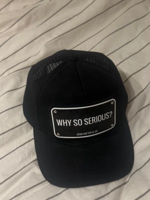 Svart keps John Hatter & Co - Snygg svart keps från John Hatter & Co med texten 'WHY SO SERIOUS?' framtill. Klassisk trucker-modell med mesh på sidorna och justerbar snapback. Perfekt för dig som vill sticka ut med en cool accessoar.