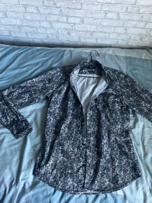 Svartvit mönstrad skjorta - Säljer en svartvit skjorta med abstrakt mönster och klassisk krage. Skjortan har långa ärmar och knäppning framtill. Materialet känns som bomull eller polyester, perfekt för dig som gillar unika prints.