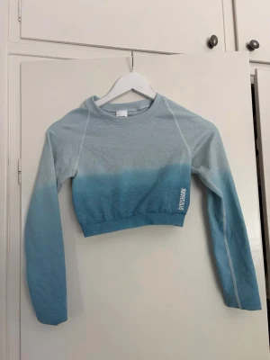 Blå ombre tränings top från Gymshark - Trendig långärmad crop top från Gymshark i blått ombre-mönster. Toppen har rund hals, vita sömdetaljer och logga på magen. Baksidan har en snygg rynkad detalj som ger extra form. Perfekt för dig som gillar sportig och modern stil.