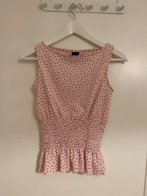 Rosa prickig ärmlös topp - Söt rosa topp med svarta prickar och smockad midja som ger en fin passform. Toppen är ärmlös och har en liten volangkant nedtill för extra detalj. Perfekt för dig som gillar en feminin och trendig stil.