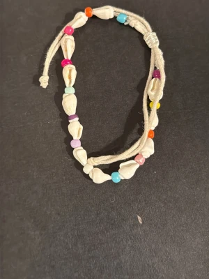 Snäckarmband med färgglada pärlor - Ett handgjort armband med vita snäckor och små färgglada pärlor i orange, rosa, blått, lila och gult. Armbandet har en justerbar knytning i beige snöre och ger en bohemisk och strandig vibe.