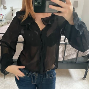 Svart transparent blus med volang - Snygg svart transparent blus med volangdetaljer vid kragen och knäppning framtill. Blusen har långa ärmar med lätt puff och är tillverkad i ett tunt, skönt material som ger en luftig känsla. Perfekt att styla med jeans eller kjol för en trendig look.