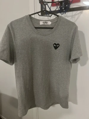Grå t-shirt med svart hjärtdetalj - Snygg grå t-shirt med rund hals och korta ärmar. På bröstet sitter en svart hjärtdetalj med ögon, vilket ger en unik touch. Tillverkad i mjuk bomull som känns skön mot huden. Perfekt för dig som gillar stilrena plagg med en twist.