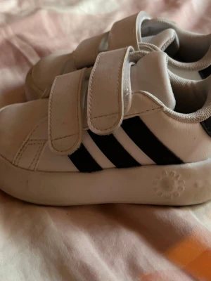 Vita sneakers med svarta ränder - Snygga vita sneakers med två kardborreband och svarta ränder på sidan. Skorna har en låg profil och är tillverkade i syntetmaterial med vadderad insida för extra komfort. Perfekta för en sportig och avslappnad stil.