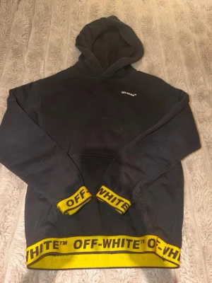 Svart hoodie från Off-White med huva - Svart hoodie från Off-White med huva och tydliga Off-White-loggor i gult på muddar och nederkant. Klassisk Off-White-etikett på bröstet. Tillverkad i mjukt bomullsmaterial och har en avslappnad passform. Perfekt för dig som gillar streetwear och vill sticka ut.
