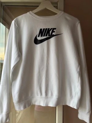 Vit sweatshirt från Nike - Säljer en clean vit sweatshirt från Nike med den klassiska svarta loggan tryckt på bröstet. Tröjan har rund halsringning och långa ärmar. Perfekt till streetwear-looken och enkel att matcha med jeans eller joggers.