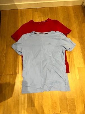 Två Tommy Hilfiger t-shirts - Säljer två klassiska t-shirts från Tommy Hilfiger. En ljusblå och en röd, båda med liten logga på bröstet. T-shirtsen har rund hals och korta ärmar, tillverkade i mjuk bomull. Perfekta till jeans eller shorts för en enkel och stilren look.